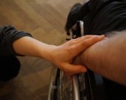 Hände zweier Menschen berühren sich am Rad eines Rollstuhls.