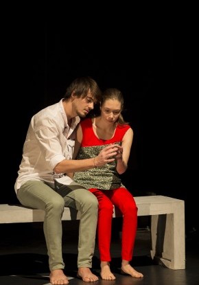 Bühnenaufnahme. Ein junger Mann und eine junge Frau sitzen nebeneinander auf einer Bank. Ihre Fingerkuppen berühren sich.