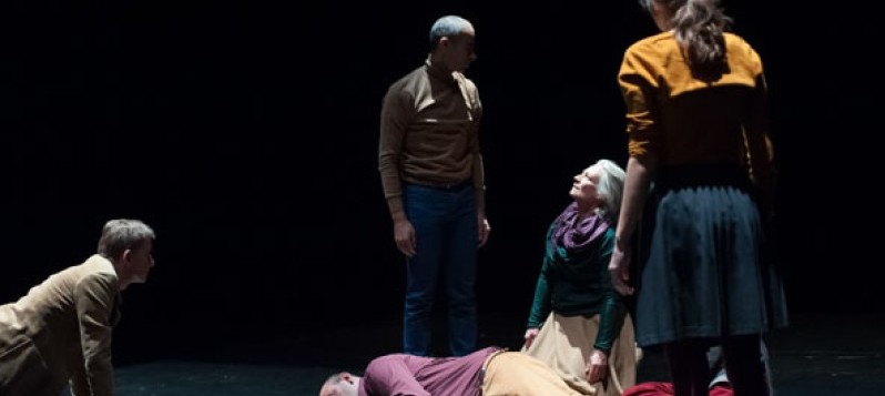 Bühnenaufnahme. Fünf Darsteller scharen sich um einen seitlich am Boden liegenden, bullig gebauten Mann.