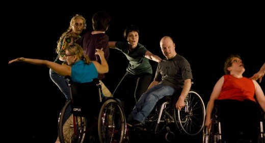 Fünf Tanzende sind kettenförmig miteinander verbunden. Eine Rollstuhlfahrerin sitzt abseits von ihnen.