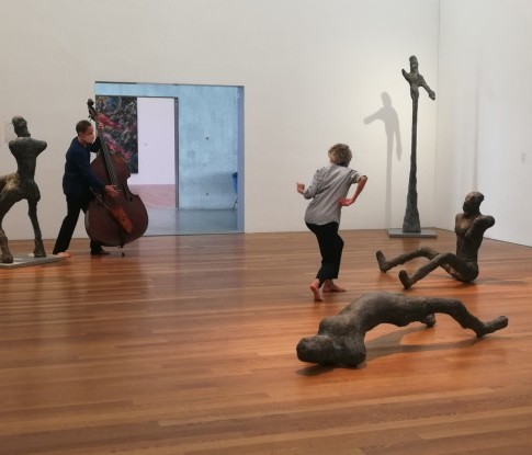 In der Mitte eines modernen Galerieraumes steht eine Tänzerin mit im Laufe gebogenen Knien und wringt ihren Körper mit asymmetrisch angewinkelten Armen nach hinten. Links hinten spielt ein Kontrabassist. Um sie verteilt sind vier liegende, sitzende oder s
