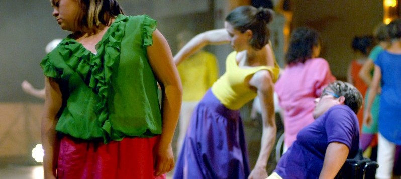 Die Performance Gruppe tanzt das Stück Sinfolie auf einer Bühne. Alle tragen unterschiedlichste Kostüme in kontrastreichen Farben. Ganz vorn blickt eine junge Frau mit Trisomie 21 in einer grünen Rüschenbluse zur Seite.