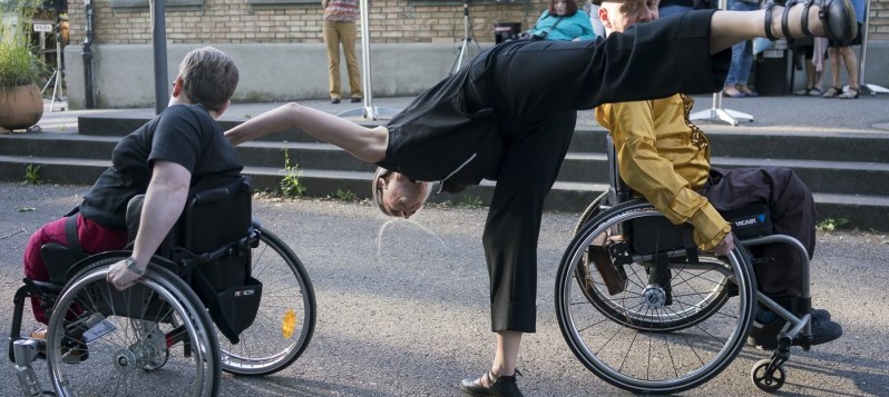Auf der Strasse hält sich eine Tänzerin tief gebeugt auf einem Bein zwischen zwei Performer:innen im Rollstuhl. 