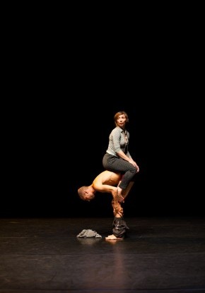 Ein Tänzer steht gebückt, barfuss auf der Bühne, eine Tänzerin sitzt auf seinem unteren Rücken.
