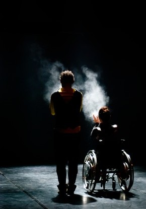 Bühnenaufnahme. Starkes Rücklicht umreisst die Silhouette eines abgewandt stehenden Mannes und einer Frau im Rollstuhl.