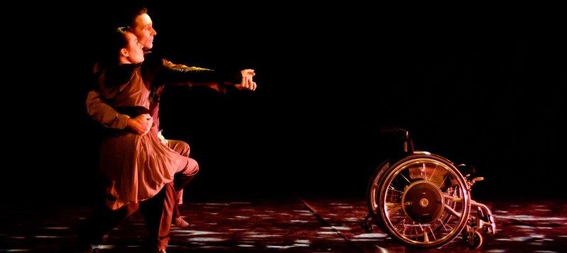 Bühnenaufnahme. Ein Paar steht im Profil in einer Tangoposition. Rechts von ihnen steht ein leerer Rollstuhl.