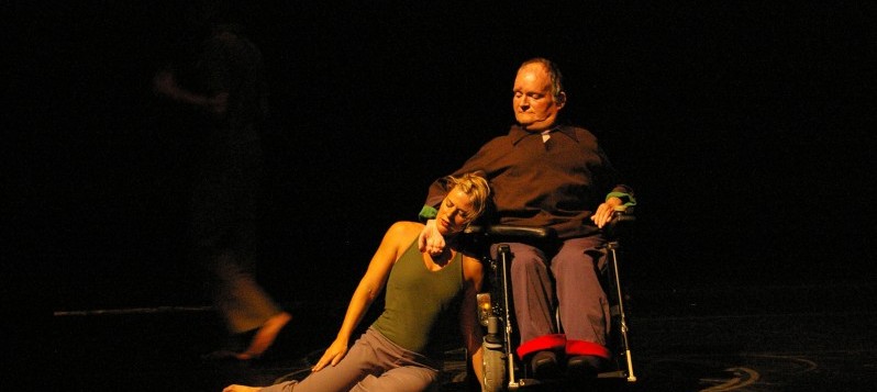 Eine Tänzerin sitzt auf dem Boden und lehnt sich an den Tänzer im Rollstuhl.