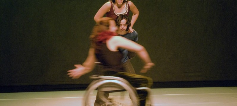 Bühnenaufnahme. Eine Frau mit schwarzem Muskelshirt im Rollstuhl fährt nach rechts vorbei. Hinter ihr stemmt sich eine Frau auf den Schultern eines Mannes in die Höhe. Ihr Mund ist schreiend aufgerissen.