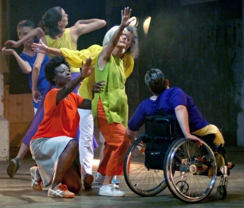 Zwei Frauen tanzen leicht versetzt nebeneinander vor einer erhöhten Bühne, auf der ein Orchester spielt. Die Tänzerinnen, eine links im Rollstuhl sitzend, die andere breit stehend, lehnen voneinander weg.