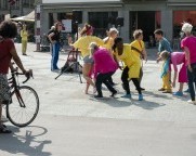 Auf der Strasse hält sich eine Tänzerin tief gebeugt auf einem Bein zwischen zwei Performer:innen im Rollstuhl. 