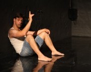 Ein Mann sitzt aufrecht auf der Bühnen. Die Beine sind angewinkelt. Der linke Arm ruht auf dem linken Knie. Der rechte Arm ist leicht angewinkelt und nach vorne ausgestreckt. Der Blick des Tänzers ist zum Publikum gerichtet.