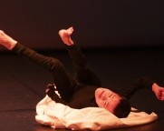 Eine Person mit kurzen schwarzen Haaren und zierlichem Körper in schwarzer Kleidung liegt auf einer Bühne, umgeben von einem weissen Stoff. Die Beine sind in die Luft gestreckt, während die Arme nach unten zeigen. 