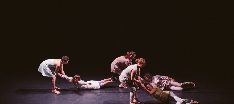 Sechs Tänzerinnen befinden sich auf einer Bühne, drei davon ziehen die anderen drei an den Armen über den schwarzen Tanzboden.