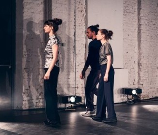 Zwei Tänzerinnen und ein Tänzer stehen am linken Rand der Bühne dicht beieinander und schauen nach vorne.