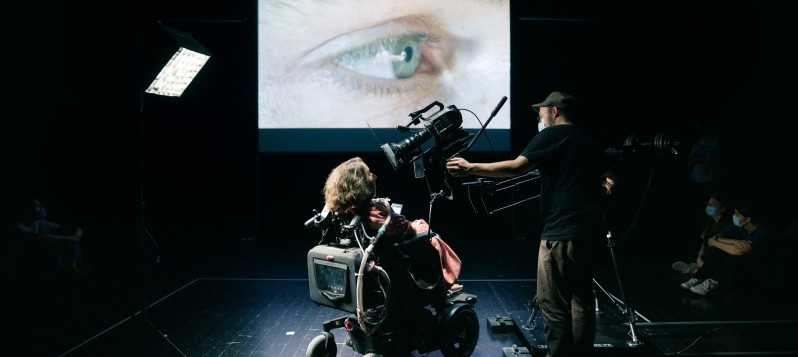 Bühnenaufnahme des Stückes «Bewegt». Eine Frau in einem Elektrorollstuhl schaut direkt in eine Filmkamera. Die Aufnahme ihres grün-blauen Auges wird auf eine Leinwand hinter ihr projiziert. 