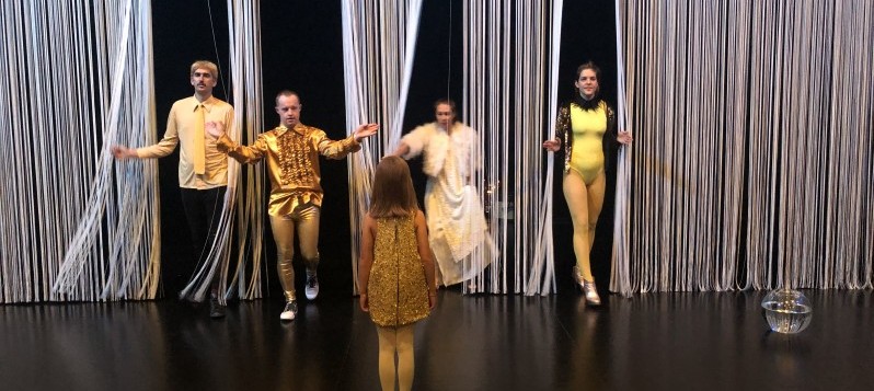 Je zwei Männer und Frauen in ausgefallenen Kostümen in Gold- und Gelbtönen schreiten durch einen weissen Faden-Vorhang auf ein Mädchen zu, das ihnen auf der vorderen Bühne gegenübersteht. Rechts steht ein mit Wasser gefülltes Goldfischglas auf dem schwarz