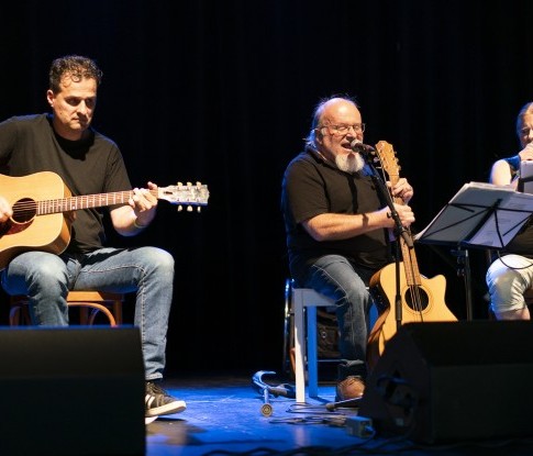 Drei Musiker:innen spielen auf der Bühne sitzend Gitarre begleitet von Gesang. 