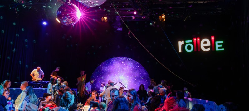 Viele fr&ouml;hliche, tanzende Menschen mit Rollschuhen oder im Rollstuhl auf der Discofl&auml;che. Etwas erh&ouml;ht steht ein DJ an einem in DJ-Pult. An der Decke strahlt eine Discokugel und im Hintergrund leuchtet in Leuchtschrift "r&ouml;llele".  