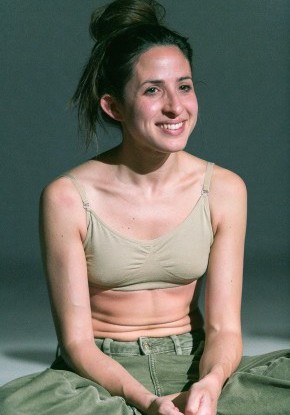 Eine Frau sitzt entspannt im Schneidersitz auf dem Boden, l&auml;chelt leicht und blickt nach vorne.