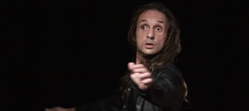 Ein Mann steht vor dunklem Hintergrund, streckt einen Arm nach vorne und schaut mit weit ge&ouml;ffneten Augen zur Seite.