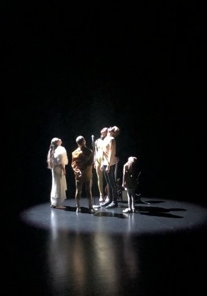 Ein Quartett mit drei Männern und einer Frau unterschiedlichen Alters tanzt dynamisch verteilt im schwarzen Bühnenraum. Alle tragen unterschiedliche Oberteile und Hosen in Rot- und Grautönen. 
